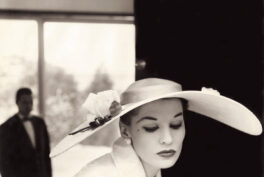 Black and white photo of a woman looking down with a big hat and with a man in the background looking at her.